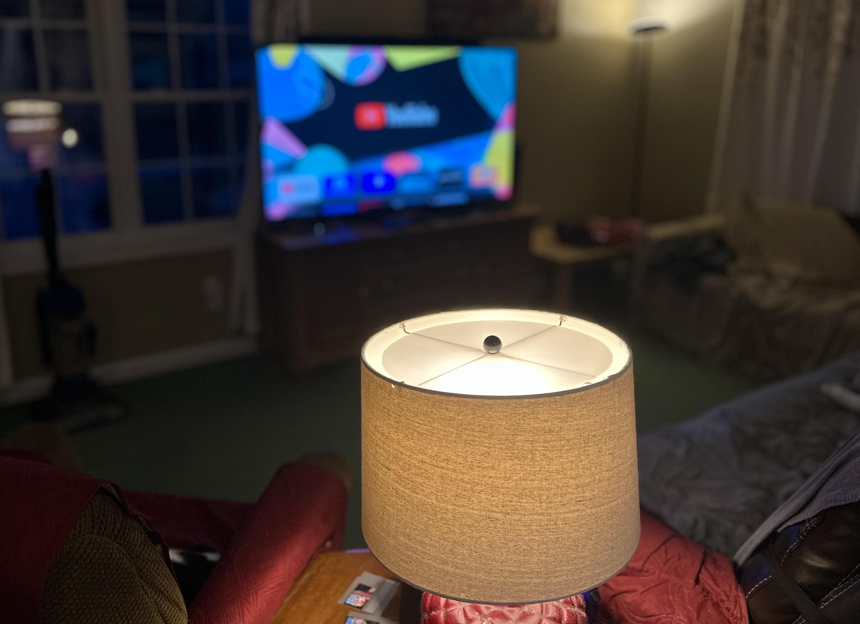 a photo of a lamp illuminated in a darkish room with a tv beyond it.