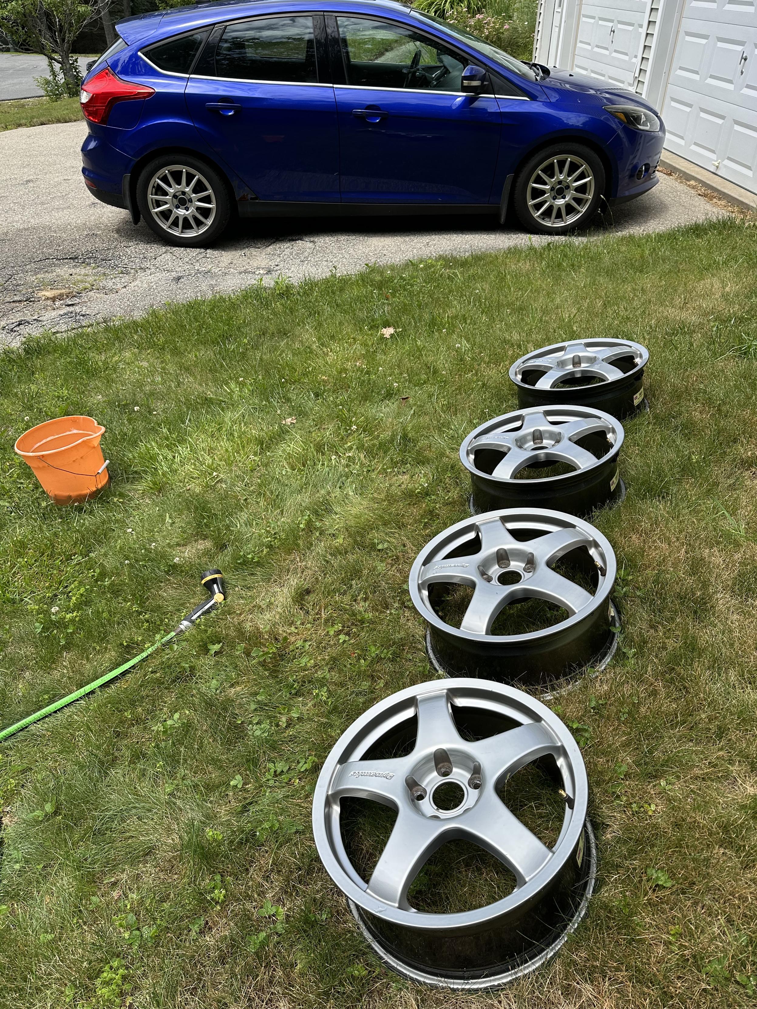 a photo of a set of car wheels in the grass, with a blue Ford Focus in the driveway beyond them. 